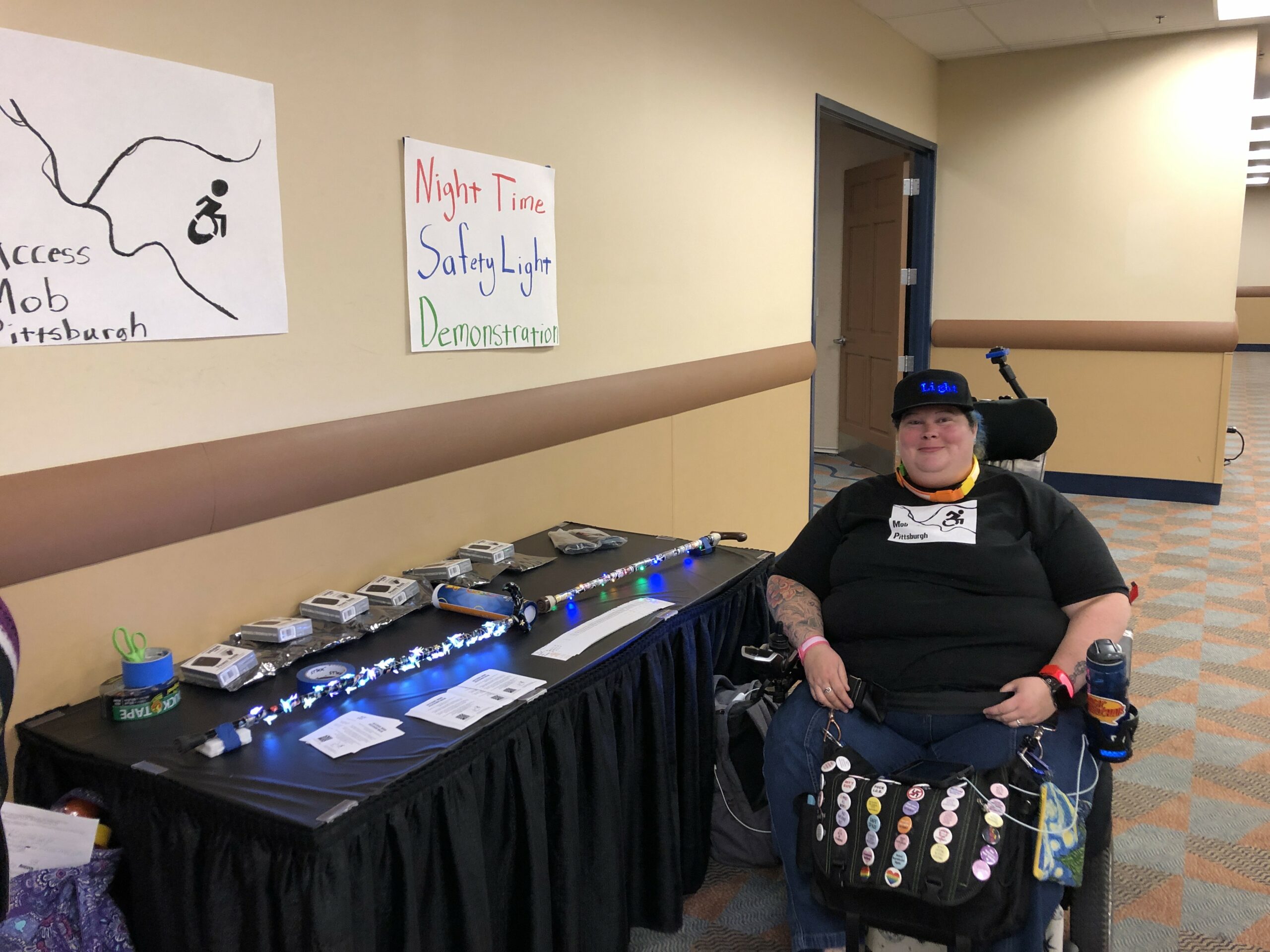 Alisa Grishman seated in her power chair next to a table that features illuminated canes.  A sign for Access Mob Pittsburgh and a sign for Night Time Safety Light Demonstration hang on the wall above the table.