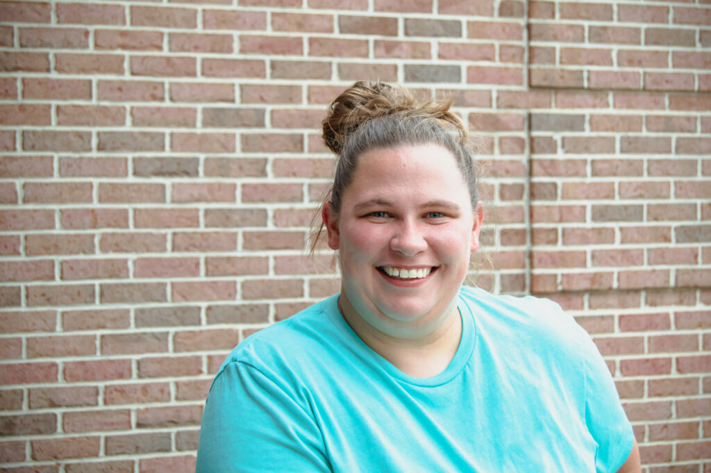 White woman smiling with hair in a messy bun, with a light blue shirt on, standing in front of a brick wall
